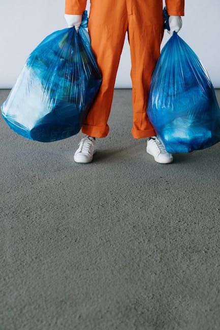 A person wearing orange work overalls and white sneakers stands on a concrete surface, holding two large blue plastic bags filled with rubbish, one in each hand. The bags are semi-transparent, reveali