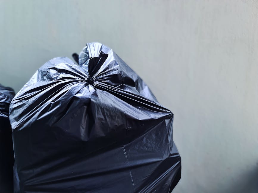 A tall, bright blue plastic rubbish bin with a closed lid, positioned outdoors on a grassy area near a sidewalk. The bin has a slightly textured surface and features a small, irregularly torn yellow s
